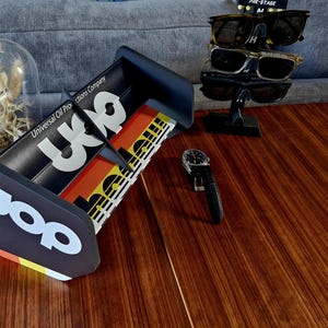 Puede incluir: Un expositor negro de UOP con texto blanco, que contiene un reloj, gafas de sol y una foto enmarcada. El expositor tiene el texto "UOP" y "Universal Oil Pro". El expositor tiene un diseño de rayas de colores.