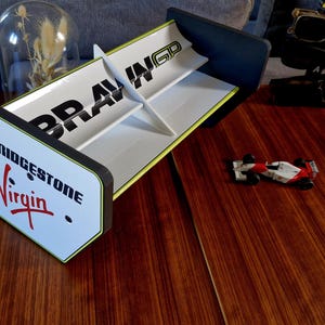 May include: A white and black Brawn GP Formula 1 rear wing with Bridgestone and Virgin logos. The wing sits on a wooden surface next to a small race car model. The words BRAWN GP are printed on the wing.