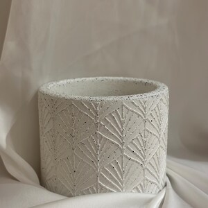 May include: A cylindrical, white planter with a textured surface and a repeating geometric leaf pattern. The planter is made of a speckled material and is suitable for indoor or outdoor use. The pot is approximately 15 cm tall.
