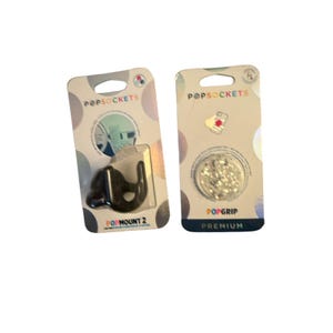 Op de afbeelding: Twee verpakte PopSockets-artikelen. Links een zwarte PopMount 2 autohouder. Rechts een ronde PopGrip Premium met een zilverkleurig sterrenpatroon. Beide artikelen zitten in de originele kartonnen winkelverpakking met merknaam.