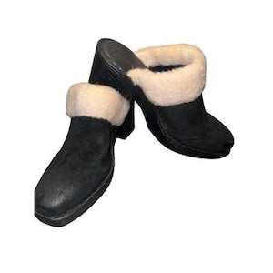 May include: A pair of black suede clogs with a cream-colored, fluffy trim around the opening. The clogs have a block heel and a rounded toe. The shoes are angled towards each other, showcasing their design.