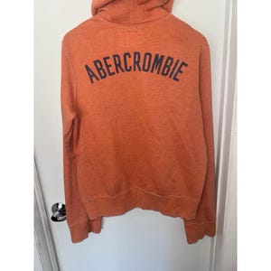 May include: An orange hooded sweatshirt featuring the word ABERCROMBIE printed in dark navy blue letters across the upper back. The garment includes a hood, long sleeves, and ribbed cuffs and hem, constructed from a soft, textured fabric.