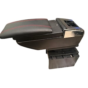 May include: A black car armrest console featuring a padded top lid with red stitching, a smooth black plastic body, and a metallic silver accent piece at the rear. The base consists of a sturdy, rectangular black plastic mounting bracket.
