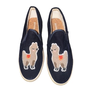 Puede incluir: Un par de zapatos sin cordones azul marino con un parche bordado de llama beige esponjosa en la parte delantera de cada zapato. Las llamas tienen mejillas rosadas y una pequeña silla de montar roja y naranja. Las plantillas son de corcho beige con texto Soludos.