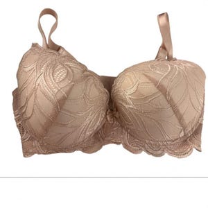 May include: A beige underwire bra featuring a textured lace overlay with a swirling leaf-like pattern, scalloped edges along the bottom band, adjustable shoulder straps, and a small decorative bow centered between the two molded cups.
