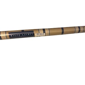 May include: A roll of Ralph Lauren Home wallpaper featuring a tan background with a repeating pattern of thin, dark vertical stripes. A black label displays the text RALPH LAUREN HOME and a smaller white label reads ORD: RED AS.