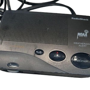 Peut inclure: Un appareil RadioShack NOAA Weatheradio Alert. L'appareil est gris foncé avec une zone de haut-parleur perforée et plusieurs boutons, dont "TEST", "ALERT" et "WEATHER". Le logo NOAA et le texte "WEATHERADIO ALERT" sont visibles.
