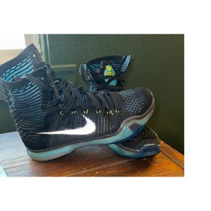 Pode incluir: Um tênis de basquete Nike de cano alto com cabedal em malha preta com detalhes horizontais azuis, um logotipo swoosh branco na lateral, cadarços pretos e uma sola azul translúcida. Um pequeno emblema circular amarelo e azul é visível.