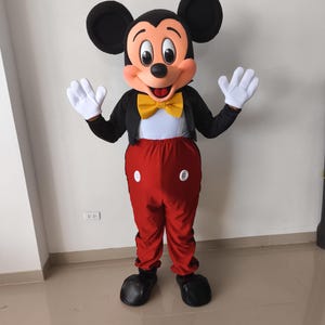 Könnte beinhalten: Ein Ganzkörper-Mickey-Mouse-Kostüm. Das Kostüm hat einen großen Kopf mit schwarzen Ohren, ein rotes und weißes Gesicht und einen schwarzen Körper mit roter Hose. Das Kostüm beinhaltet weiße Handschuhe, schwarze Schuhe und eine gelbe Fliege.