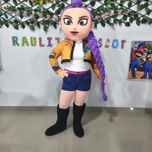 Könnte beinhalten: Ein Maskottchen-Kostüm einer Zeichentrickfigur mit großem Kopf, lila Haaren und einer gelben Jacke. Das Kostüm umfasst ein weißes Hemd, blaue Shorts und schwarze Stiefel. Der Hintergrund zeigt den Text "RAULITO SCOT".