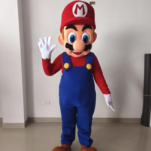 Könnte beinhalten: Ein Ganzkörperkostüm, das der Figur Mario ähnelt. Das Kostüm hat einen roten Hut mit einem weißen "M", ein rotes Langarmshirt, eine blaue Latzhose und große braune Schuhe. Weiße Handschuhe bedecken die Hände, und die Figur winkt.