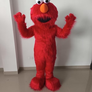 Könnte beinhalten: Ein rotes Ganzkörperkostüm, das einer beliebten Kinderfigur ähnelt. Das Kostüm hat große weiße Augen, eine orangefarbene Nase und einen breiten, lächelnden Mund. Das Plüschmaterial bedeckt den gesamten Körper, einschließlich großer Füße und Hände.