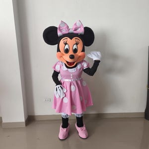 Könnte beinhalten: Ein Minnie Mouse Kostüm mit einem rosa Kleid mit weißen Punkten, einer passenden Schleife und schwarzen Ohren. Das Kostüm umfasst weiße Handschuhe, schwarze Ärmel und rosa Schuhe. Die Figur winkt.