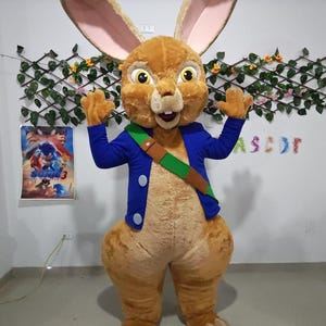 Könnte beinhalten: Ein Ganzkörper-Hasen-Maskottchen-Kostüm mit braunem und beigem Fell. Das Kostüm hat große Ohren, eine blaue Jacke und eine grüne Schärpe. Das Maskottchen steht mit erhobenen Armen, im Hintergrund ist das Wort "ASCOT" zu sehen.
