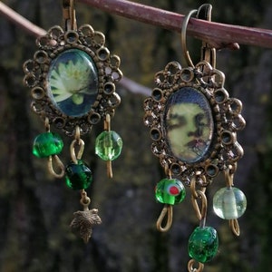 May include: A pair of antique-style earrings with oval-shaped pendants featuring botanical and portrait images. The earrings have a bronze-colored filigree frame, green glass beads, and small leaf charms. The earrings are hanging from a tree branch.