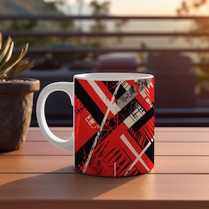 May include: White ceramic mug with a bold geometric design in red, black, and white. The pattern features intersecting lines and shapes, creating a modern, abstract aesthetic. The mug has a standard handle and is set on a wooden surface.