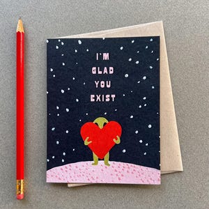 May include: A greeting card with a black background and white polka dots. A green alien figure is holding a red heart. The text on the card reads "I'm Glad You Exist."