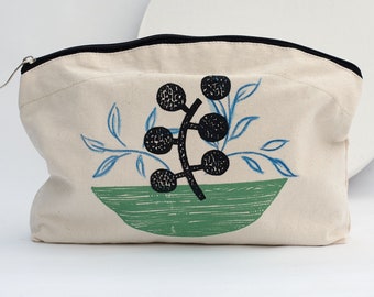 Könnte beinhalten: Eine cremefarbene Canvas-Reißverschlusstasche mit einem schwarzen Reißverschluss und einem schwarzen und grünen Aufdruck einer stilisierten Pflanze mit schwarzen Beeren in einer grünen Schüssel.