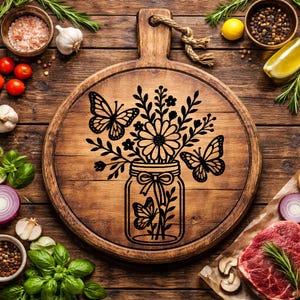 May include: A round wooden cutting board with a handle and a decorative black floral and butterfly design. The design features a jar filled with flowers and butterflies. The board is surrounded by ingredients like tomatoes, garlic, and herbs.