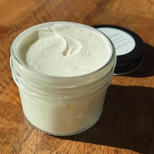 May include: A clear glass jar filled with thick white cream, featuring a smooth, swirled surface texture. Beside the jar sits a black circular lid with a white label that includes the text 'Lavender' and '4 oz' printed in a simple font.
