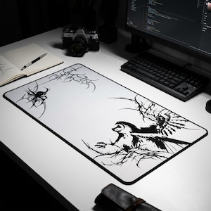 Op de afbeelding: Een grote witte muismat met een zwarte rand. De mat heeft een zwart-wit grafisch ontwerp van een vogel met uitgespreide vleugels en abstracte vormen. Een camera, toetsenbord en notitieboek staan op de achtergrond.