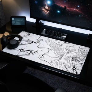 May include: A white rectangular mouse pad featuring a black line drawing of an angel with wings, stars, and flowing lines. The pad is on a black desk with a computer monitor, keyboard, and headphones.