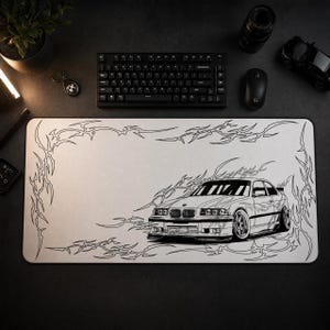 May include: A rectangular desk mat featuring a white surface with a black line art illustration of a BMW E36 coupe. The design includes intricate tribal-style flame patterns surrounding the car and framing the edges of the mat.