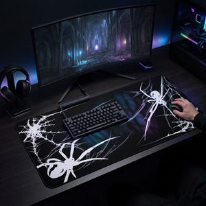 May include: Black gaming mouse pad with a white spider and web design. The pad features a keyboard, mouse, and a curved monitor displaying a digital image. A headset is also visible. The pad is designed for gaming.