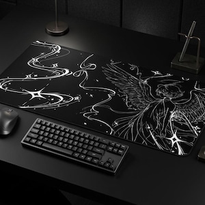 Puede incluir: Alfombrilla de escritorio en blanco y negro con un diseño de ángel y patrones de estrellas. Un teclado, un ratón y un portalápices negros también están en el escritorio. La alfombrilla es rectangular y cubre gran parte de la superficie del escritorio.