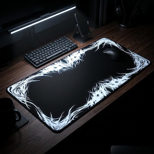May include: A black rectangular gaming mouse mat with a white, stylized, glowing border design. A black mouse rests on the mat, with a black keyboard and a phone stand visible in the background. The mat sits on a wooden desk.