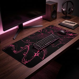 May include: Black gaming mousepad with a pink graphic design featuring a winged figure and starbursts. A black keyboard and mouse sit on the pad, alongside a computer monitor, speakers, and headphones on a wooden desk.