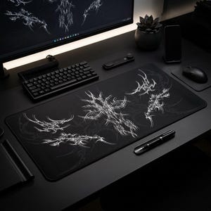 May include: Black rectangular desk mat featuring a white abstract design. The mat is surrounded by a keyboard, mouse, and other desk accessories. The design is a series of interconnected, swirling shapes.