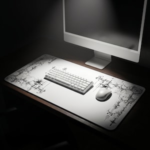 May include: A rectangular white desk mat featuring intricate black abstract line art patterns along the corners. The mat is positioned under a white mechanical keyboard and a white computer mouse, providing a clean surface for computer peripherals.