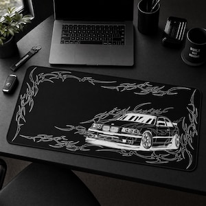 May include: A rectangular black desk mat featuring a white line art illustration of a sports car positioned in the lower right corner, surrounded by intricate tribal-style patterns that frame the edges of the mat on a dark surface.