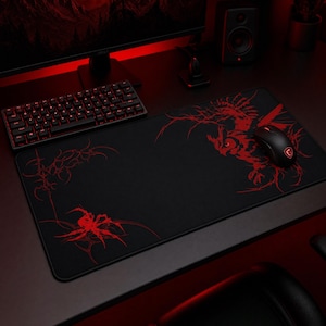 May include: A rectangular black desk mat featuring a red graphic print of an owl and a spider with intricate, web-like line work. The mat has a smooth surface with a stitched border, providing a wide area for a computer mouse and keyboard setup.