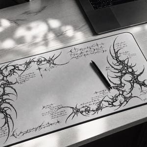 May include: A rectangular desk mat featuring a light grey surface with intricate black tribal-style line art and handwritten-style script text. A black pen rests diagonally on the mat, which is positioned on a desk next to a laptop.