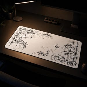 May include: A rectangular desk mat featuring a light grey surface with a black, intricate, thorn-like graphic pattern printed along the edges. The design consists of sharp, interconnected lines and cross-shaped motifs that frame the central area.