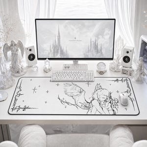 May include: A rectangular white desk mat featuring a black line art illustration of a winged figure with long hair. The design includes decorative corner accents and small star motifs. The mat is positioned on a desk with a computer monitor, keyboard, and mouse.