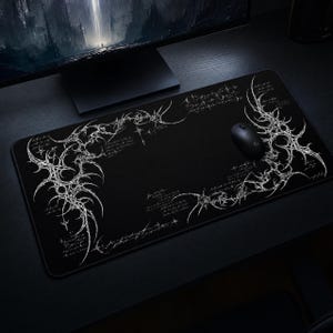 Puede incluir: Una alfombrilla de escritorio rectangular negra con intrincados diseños de líneas blancas, patrones similares a espinas y texto estilizado. Un ratón de ordenador negro descansa en el lado derecho de la alfombrilla, sobre una superficie oscura.