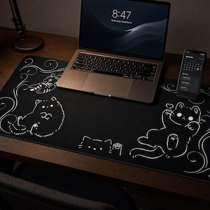 Pode incluir: Um tapete de secretária retangular preto com ilustrações de linhas brancas de três gatos em várias poses, acentuado com padrões decorativos em espiral. O tapete está posicionado sobre uma superfície de madeira escura sob um portátil.