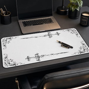 May include: A rectangular desk mat featuring a white surface with black ornate filigree corner accents and two horizontal sword illustrations centred along the top and bottom edges. A black fountain pen rests on the mat's surface.