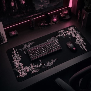 May include: A rectangular black desk mat featuring intricate, light pink gothic-style filigree patterns along the corners. A black mechanical keyboard with backlit keys and a black wireless computer mouse are positioned on the mat surface.