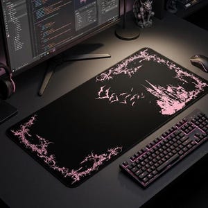 Puede incluir: Alfombrilla de ratón para juegos negra con un diseño de castillo y murciélagos en rosa, un teclado a juego y un ratón. La alfombrilla tiene un diseño de borde rosa. Un auricular negro con detalles en rosa se encuentra a la izquierda de la alfombrilla. Un monitor de ordenador está en el fondo.
