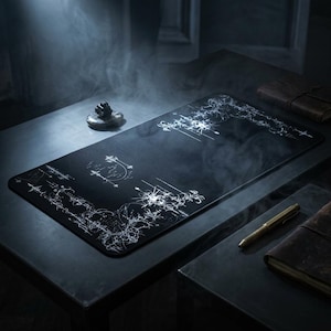 May include: A black rectangular desk mat with a white abstract design. The mat is on a dark surface, with a small sculpture and a pen nearby. The design features lines and shapes, creating a unique aesthetic.