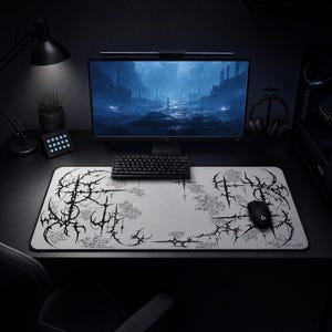 Puede incluir: Una alfombrilla de escritorio rectangular con una superficie gris claro que presenta intrincada caligrafía negra de estilo gótico y patrones abstractos similares a espinas. La alfombrilla se sitúa sobre un escritorio oscuro bajo un monitor.
