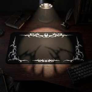 May include: Black rectangular mouse mat with a white, stylized, decorative design. The design features a central emblem and intricate patterns along the edges. The mouse mat is on a wooden desk, illuminated by a desk lamp.