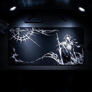 May include: Black rectangular gaming mouse pad featuring a white illustration of a knight holding a sword, with a halo and flowing cape. The design includes a cracked, web-like pattern. A black computer mouse is on the pad.