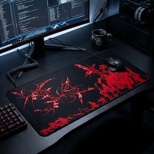 Könnte beinhalten: Schwarzes Gaming-Mauspad mit rotem, abstraktem Design. Eine schwarze Maus und eine Tastatur befinden sich auf dem Pad. Ein Computermonitor und eine Tasse mit Totenkopf-Design sind im Hintergrund. Das Pad liegt auf einem schwarzen Schreibtisch.