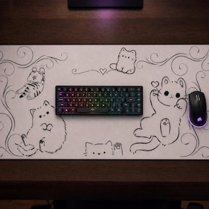 May include: A rectangular white desk mat featuring black line art illustrations of several cats in various poses, accented by swirling decorative flourishes and small hearts. A black mechanical keyboard with RGB lighting and a black computer mouse.