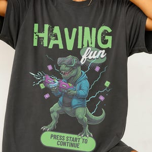 Puede incluir: Camiseta gris oscuro con un gráfico de un dinosaurio con gafas de realidad virtual y una sudadera con capucha azul, sosteniendo una pistola morada y rosa. El texto "HAVING fun" está encima del dinosaurio y "PRESS START TO CONTINUE" debajo.